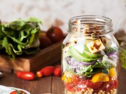 Layered salad in a jar with mixed vegetables and feta cheese, surrounded by cherry tomatoes.